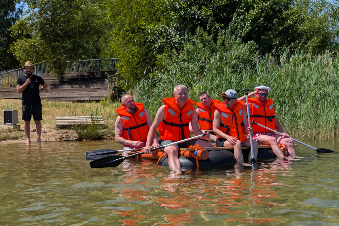 Team 2 mit Schwimmwesten und Paddeln auf ihrem Floß auf dem See mit Jakob von Teamhelden im Hintergrund